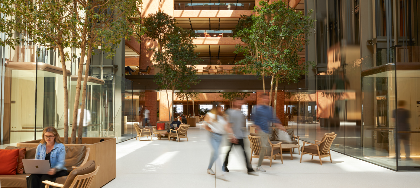 The lower floor of the light-filled Battersea atrium with multiple Apple team members meeting, walking, and working in various seating arrangements.