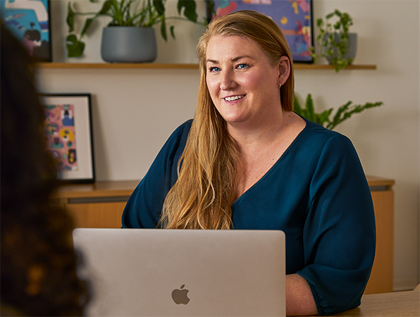 Une employée d’Apple est assiste à une table dans une aire commune et utilise un MacBook tout en parlant avec quelqu’un d’autre de l’équipe.