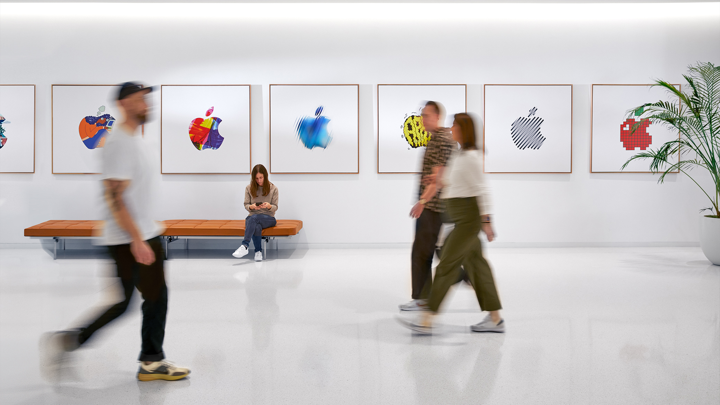 Trois membre de l’équipe Trois membres de l’équipe Apple passent devant un mur orné de logos Apple colorés, et une employée est assise sur un banc.