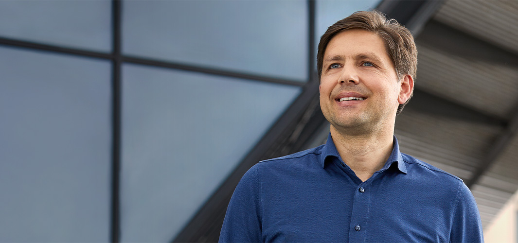 Greg standing outdoors in front of a window-paned wall, looking to the left, smiling.