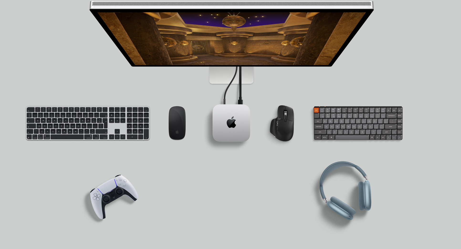 Top view of silver Mac mini in a desk setup with a display screen, wireless keyboards, wireless mice, a gaming controller, and AirPods Max