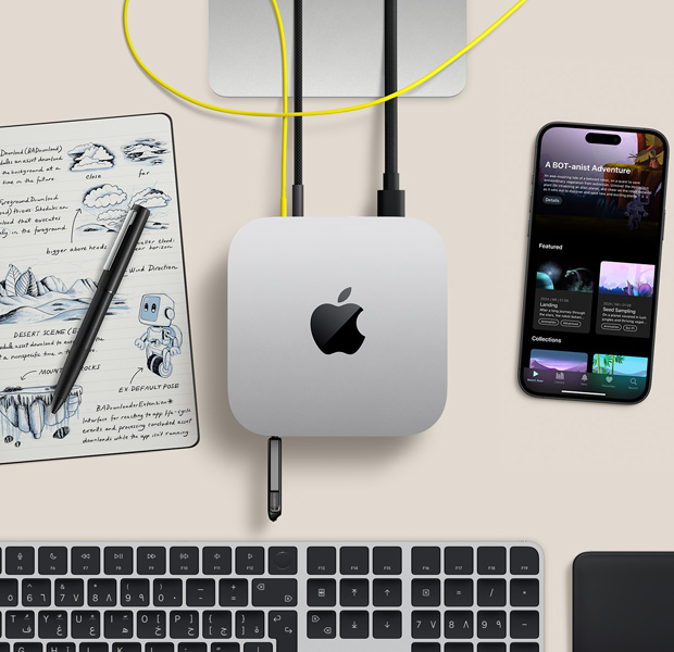 لقطة علوية لجهاز Mac mini فضي اللون مع أسلاك ومحرك أقراص محمول متصل بالمنافذ، ضمن إعداد مكتبي مع لوحة مفاتيح لاسلكية ولوحة تعقب وجهاز iPhone ودفتر ملاحظات