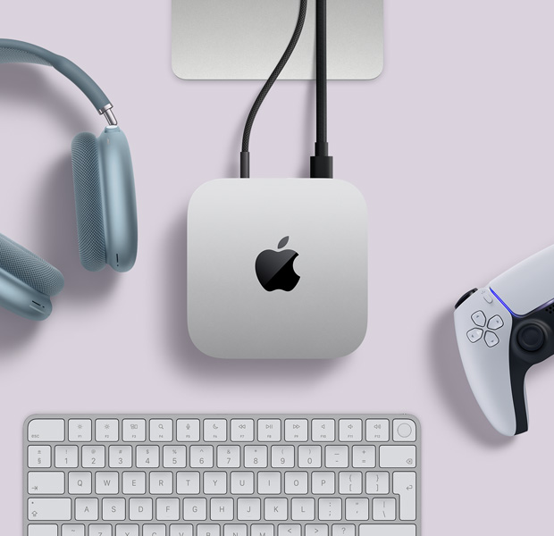 Top view of silver Mac mini, with two cords plugged into back ports, next to blue AirPods Max, a silver Magic Keyboard, and a wireless game controller