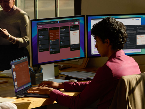 A team of employees sitting around a table in an office setting. A woman sits at her desk working on a MacBook and displays. Her colleagues are gathered around in discussion.